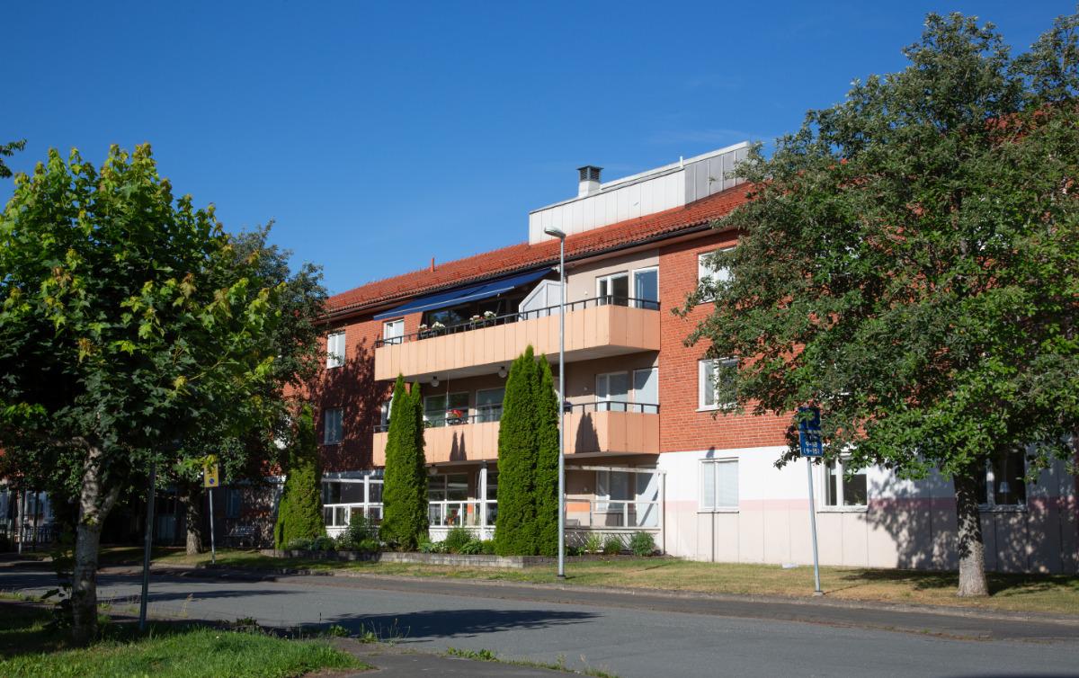 Flerbostadshus i rött tegel med balkonger, omgivet av gröna lövträd en sommardag