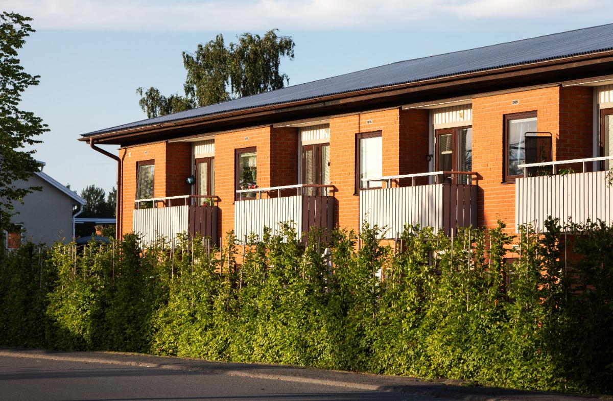 Bostadshus i rött tegel med balkonger bakom en grön häck i kvällssol